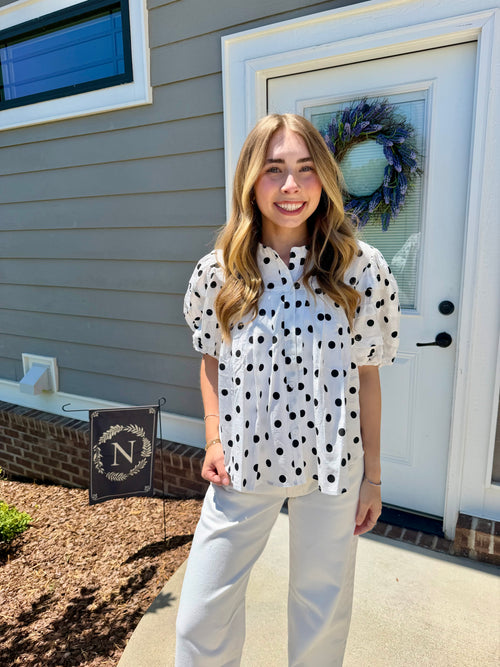 Woman in a polka dot blouse and white pants standing outside a house with a wreath on the door.