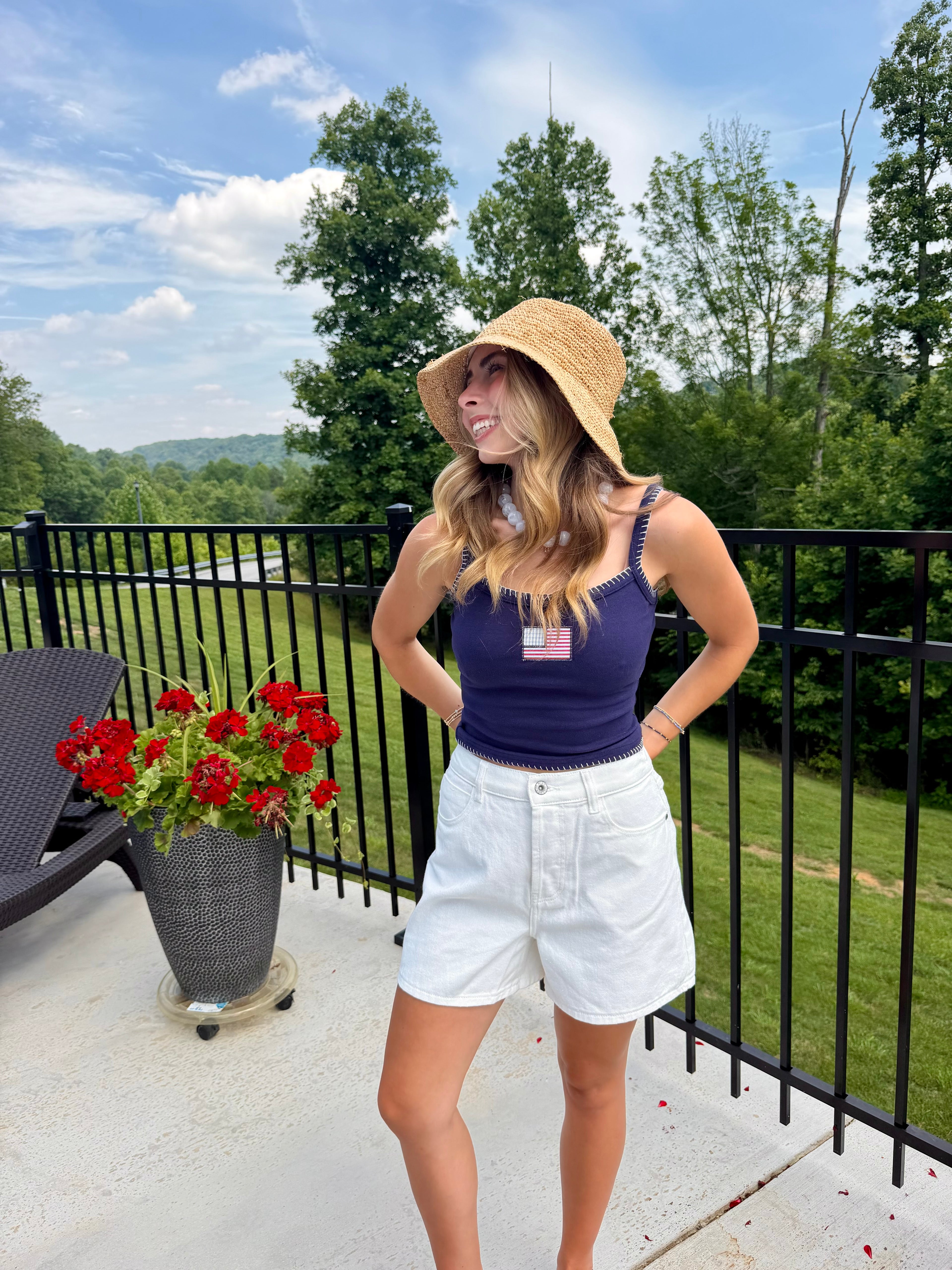 Woman on a balcony wearing a sun hat and tank top, with a scenic background of trees and sky.