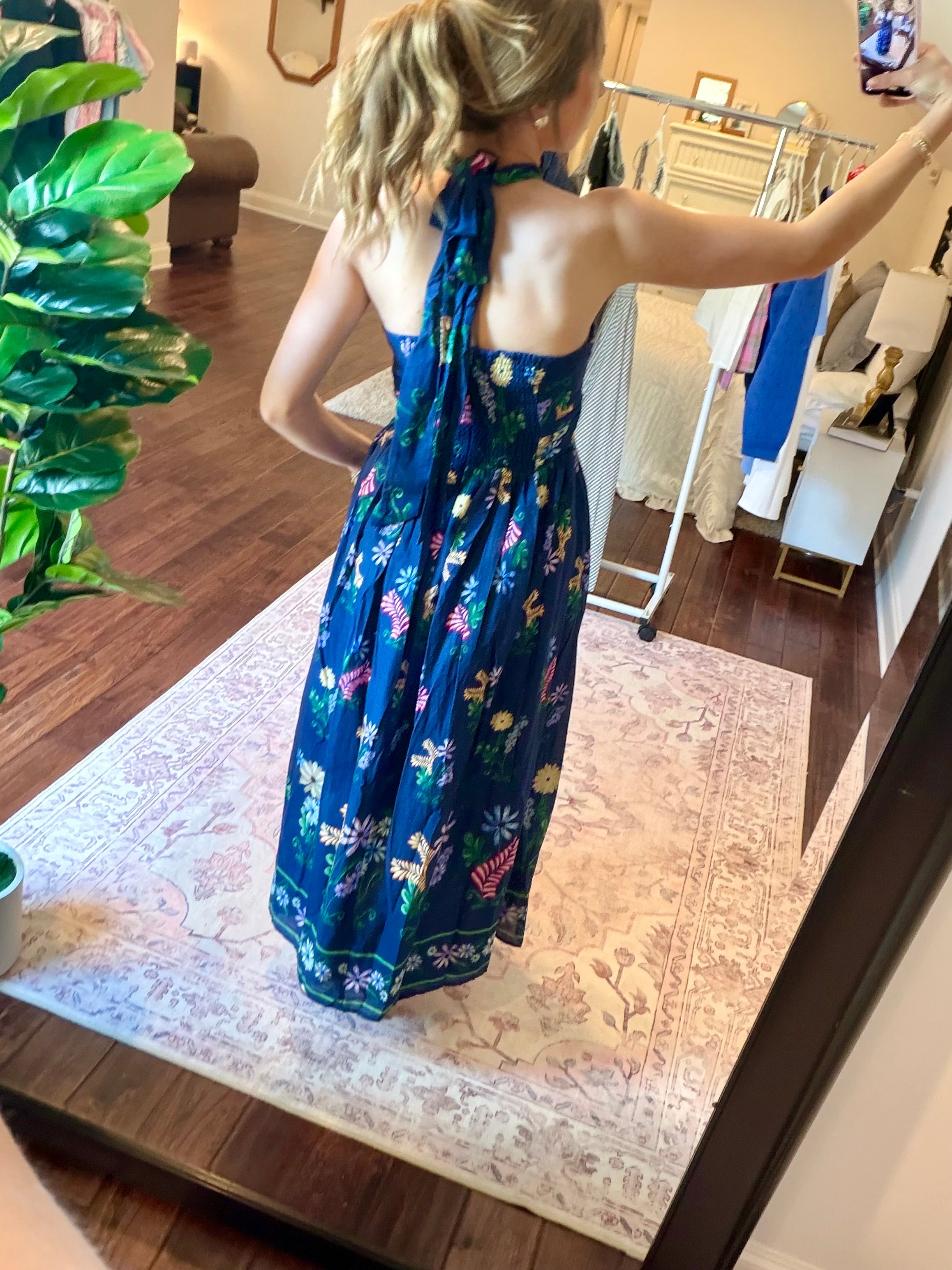 Woman taking a mirror selfie wearing a blue floral dress  in a room with a plant and decorative wall art.