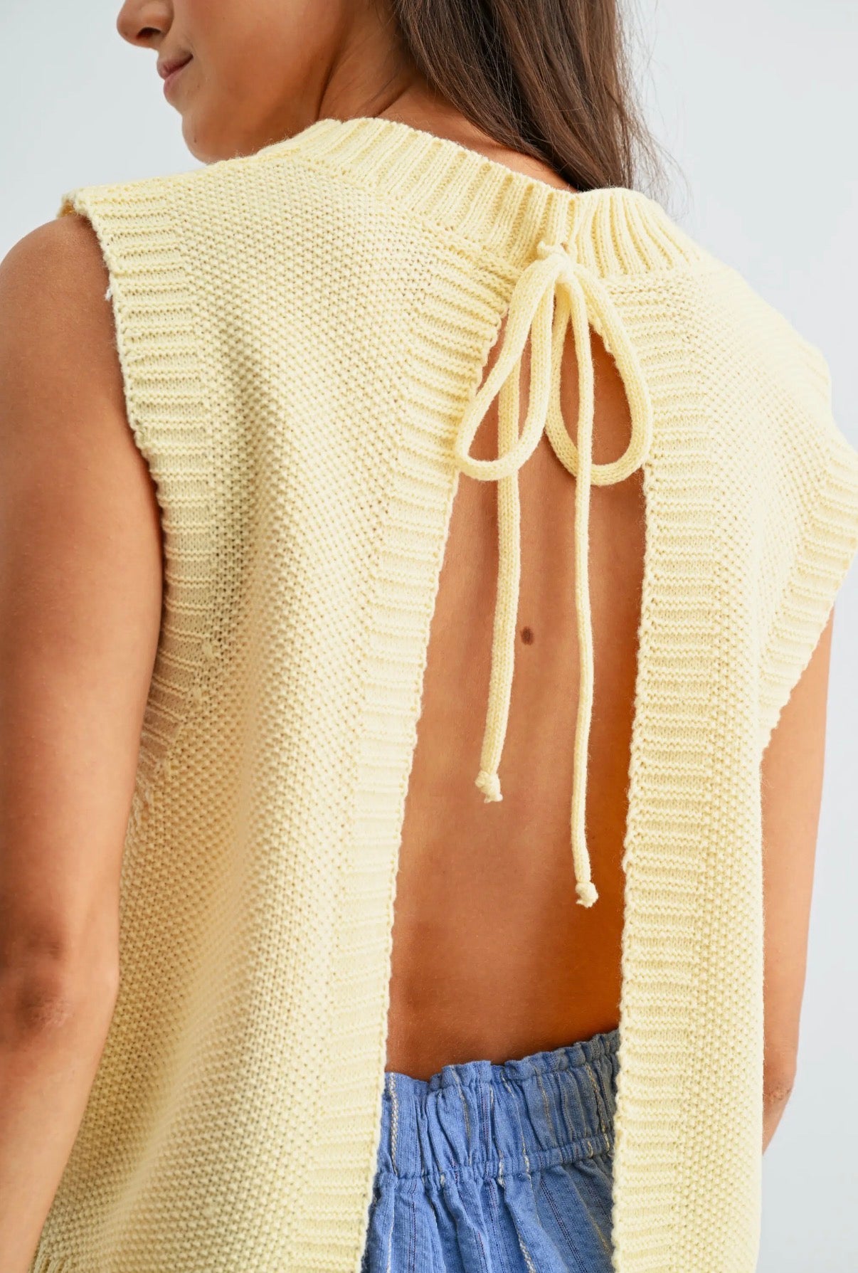 Woman wearing a light yellow knitted sleeveless top with open back, standing against a plain background.