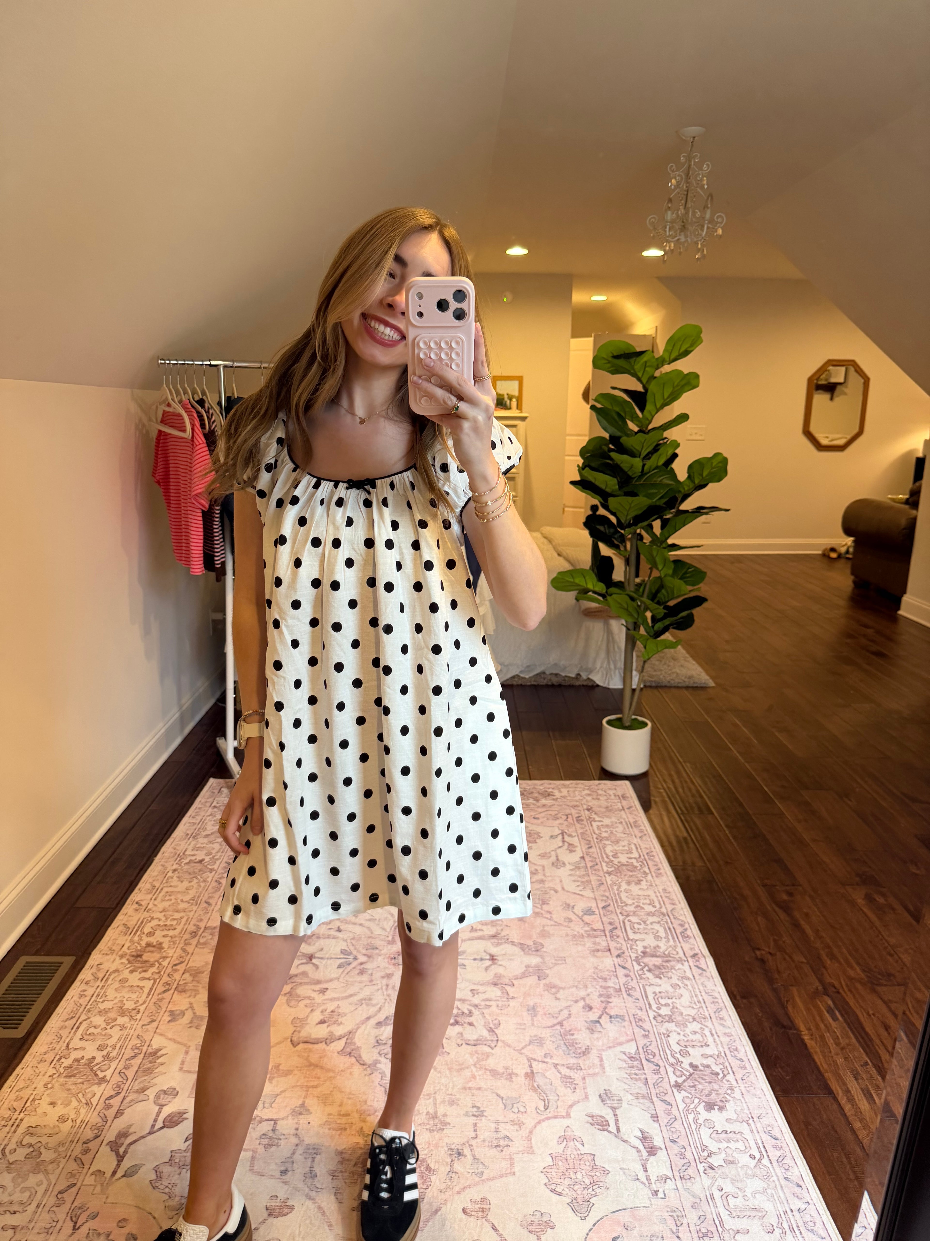 Woman in a polka dot dress standing in a room with a plant and patterned rug.