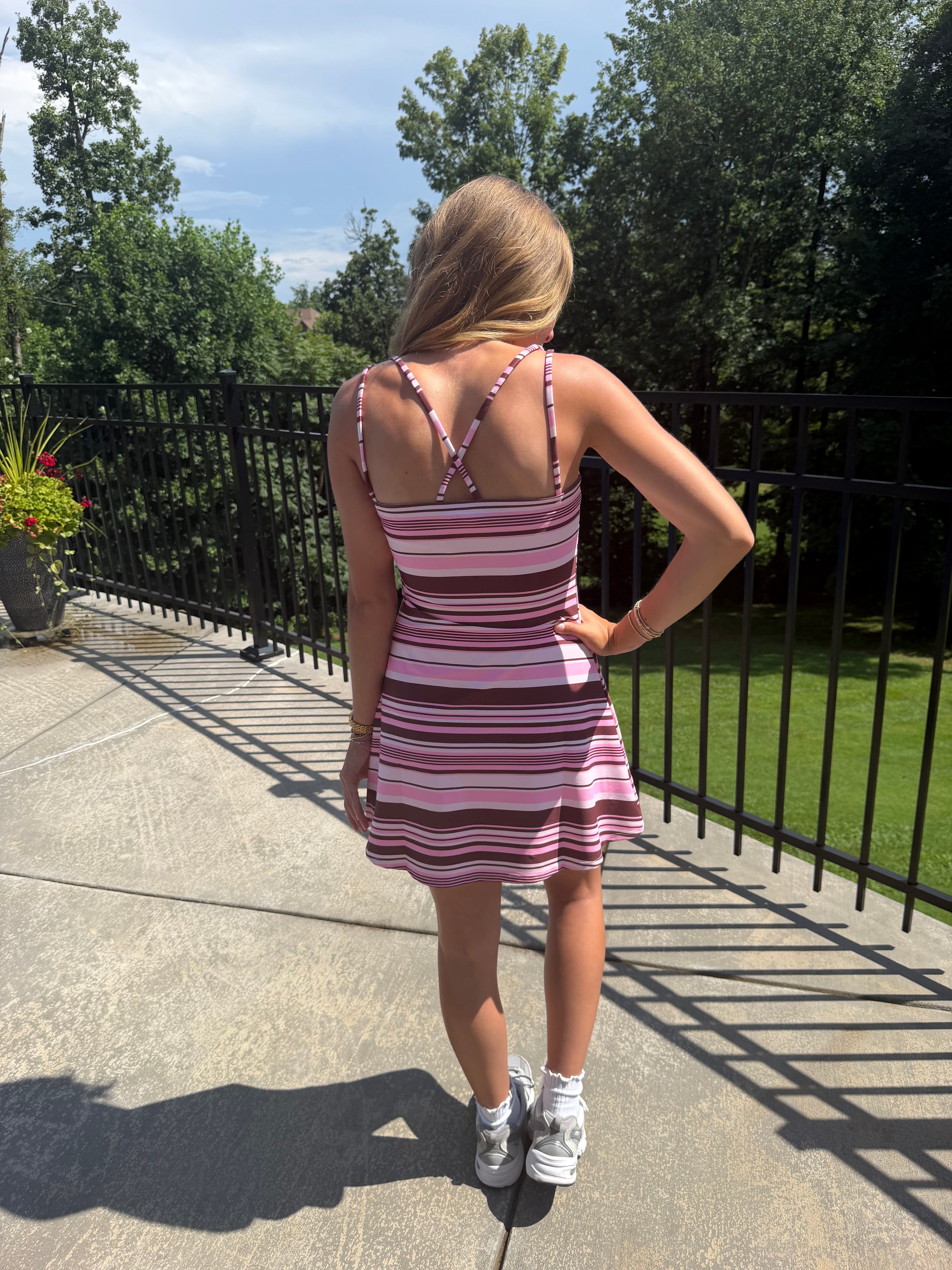 A woman stands on an outdoor patio wearing a pink and white striped mini dress with thin straps and a crisscross back design, paired with white sneakers.