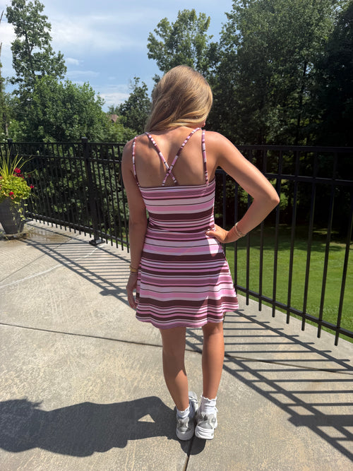 A woman stands on an outdoor patio wearing a pink and white striped mini dress with thin straps and a crisscross back design, paired with white sneakers.
