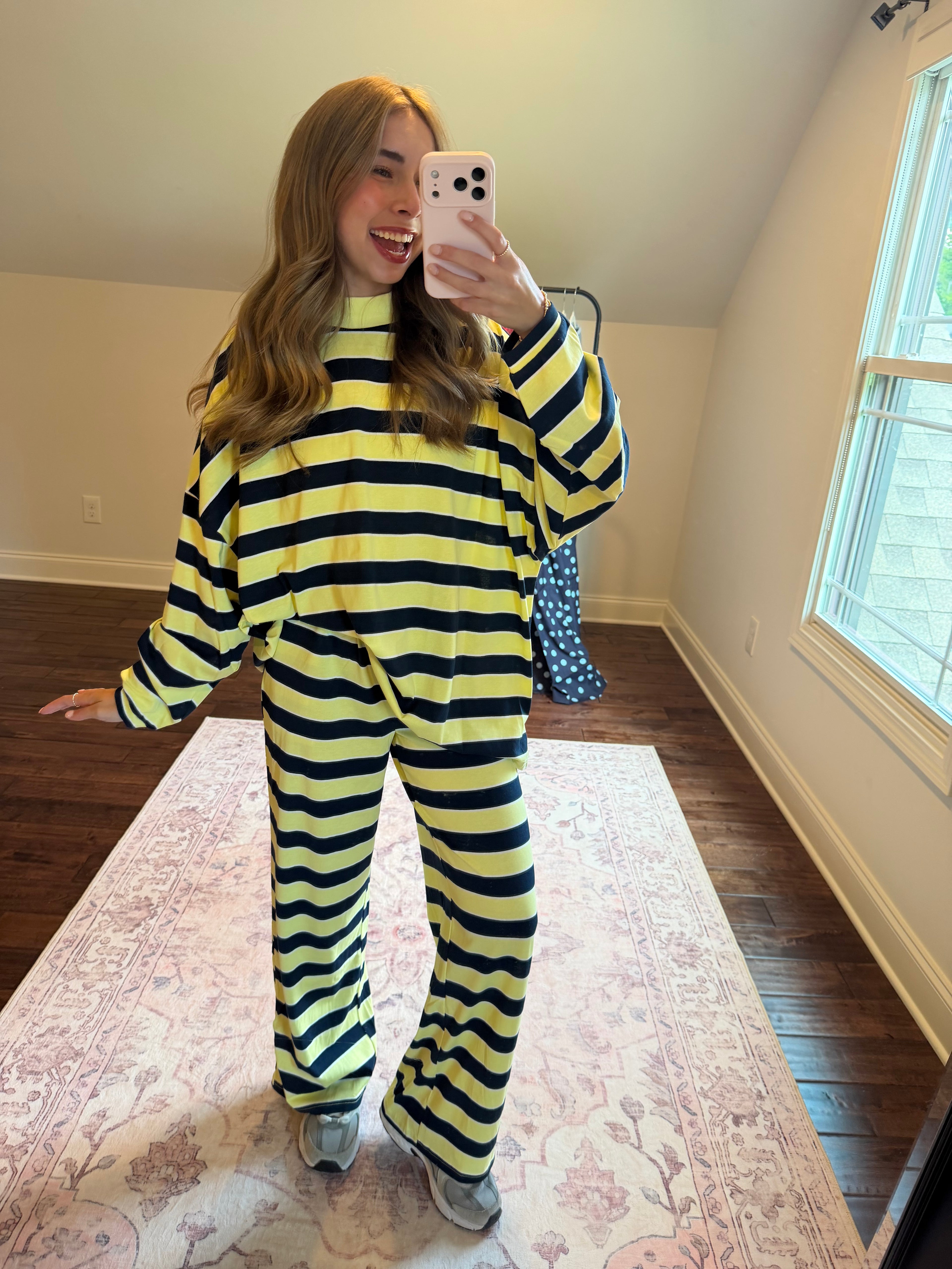 A woman takes a mirror selfie wearing a matching yellow and black striped lounge set in a room with hardwood floors and a patterned rug.