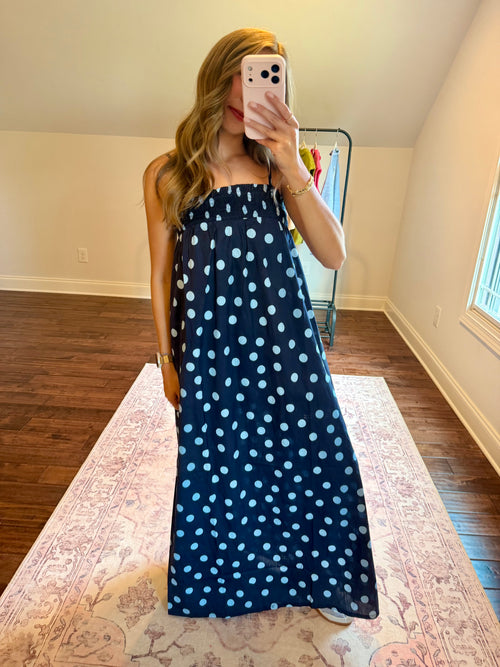 A woman takes a mirror selfie in a room wearing a navy blue strapless maxi dress with a light blue polka dot pattern. The room has a wood floor, a patterned rug, and a window.