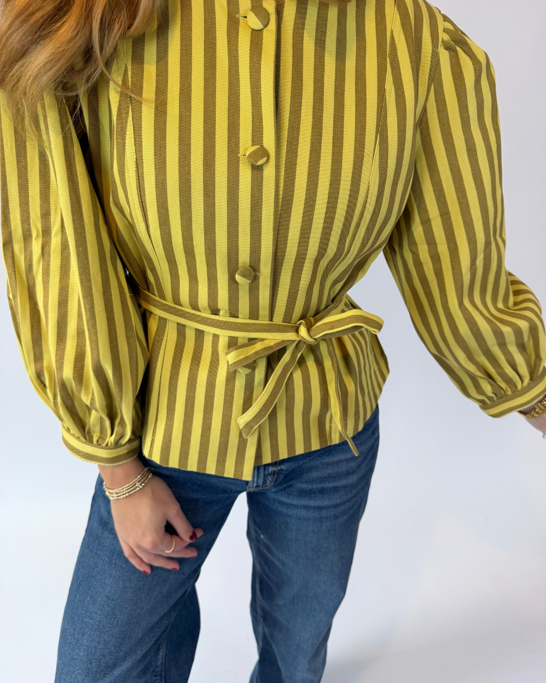 A woman wearing a yellow and dark green vertically striped long-sleeve blouse with a matching tie belt, paired with blue jeans, shown from the side against a plain white studio background.