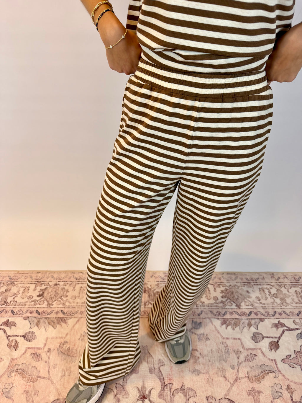 A person wearing brown and cream striped wide-leg pants and a matching striped top, standing on a patterned rug against a plain wall.