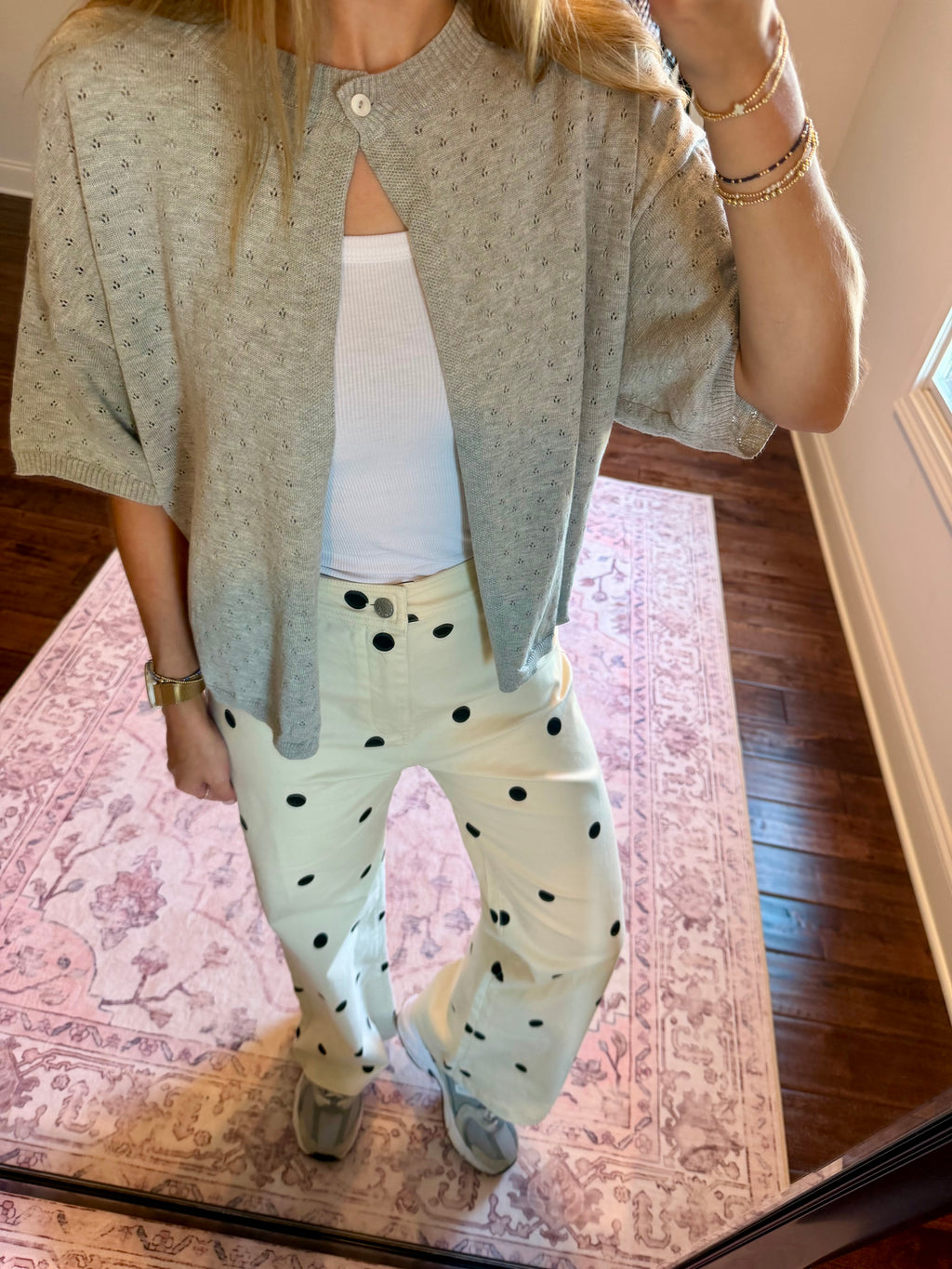 A person wearing a light gray short-sleeve knit cardigan over a white tank top and off-white wide-leg pants with black polka dots, standing indoors on a patterned rug.