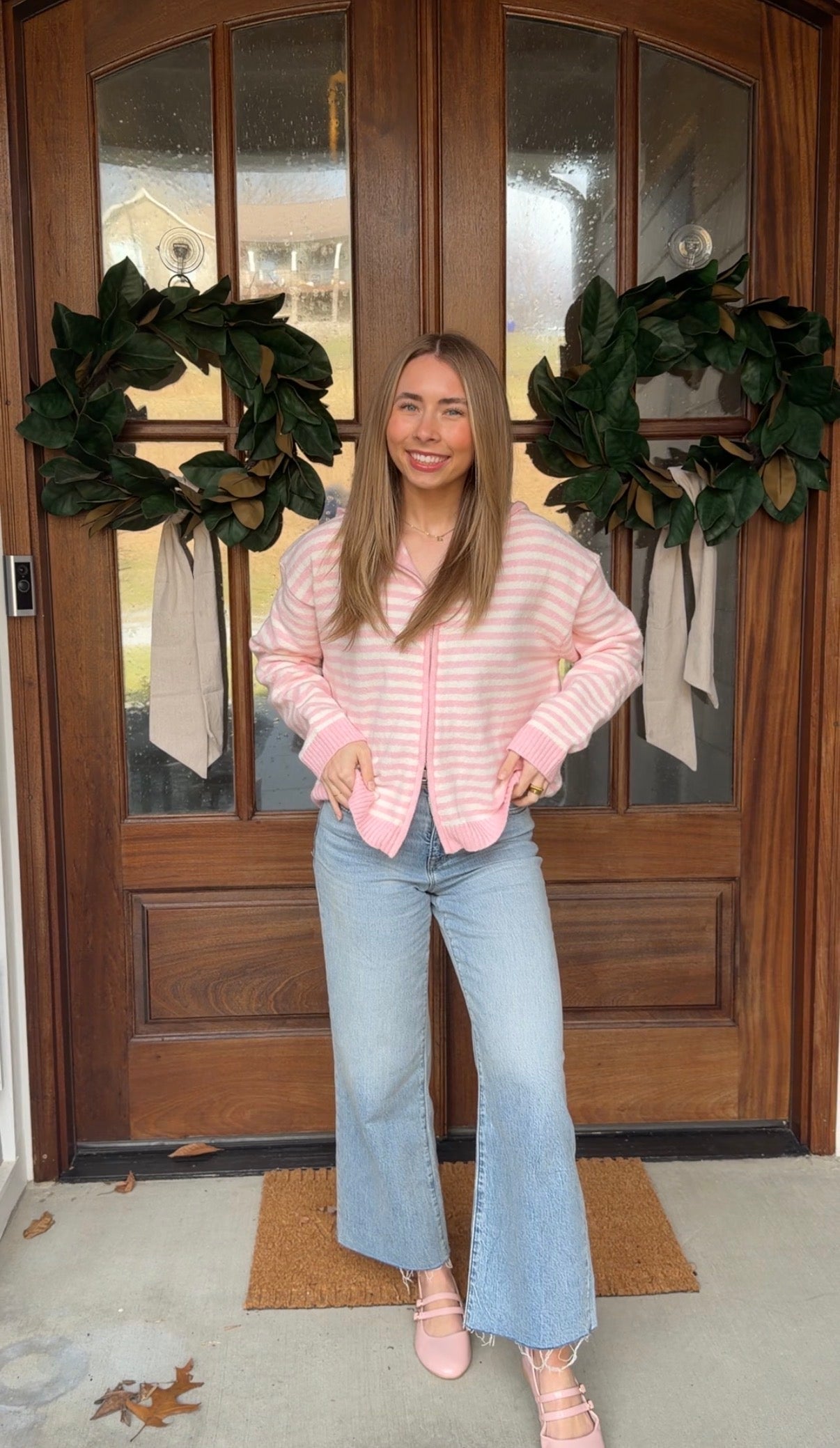 Woman standing in front of a wooden door with wreaths, wearing a pink striped sweater and light blue jeans.