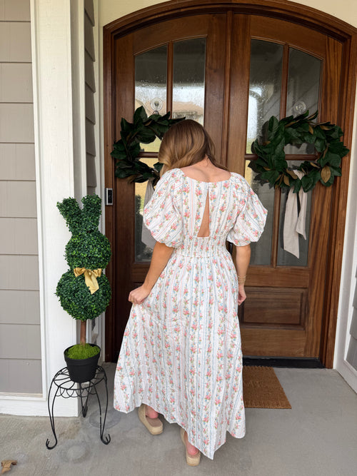 Person wearing a white dress with floral patterns standing in front of a wooden door with wreaths.