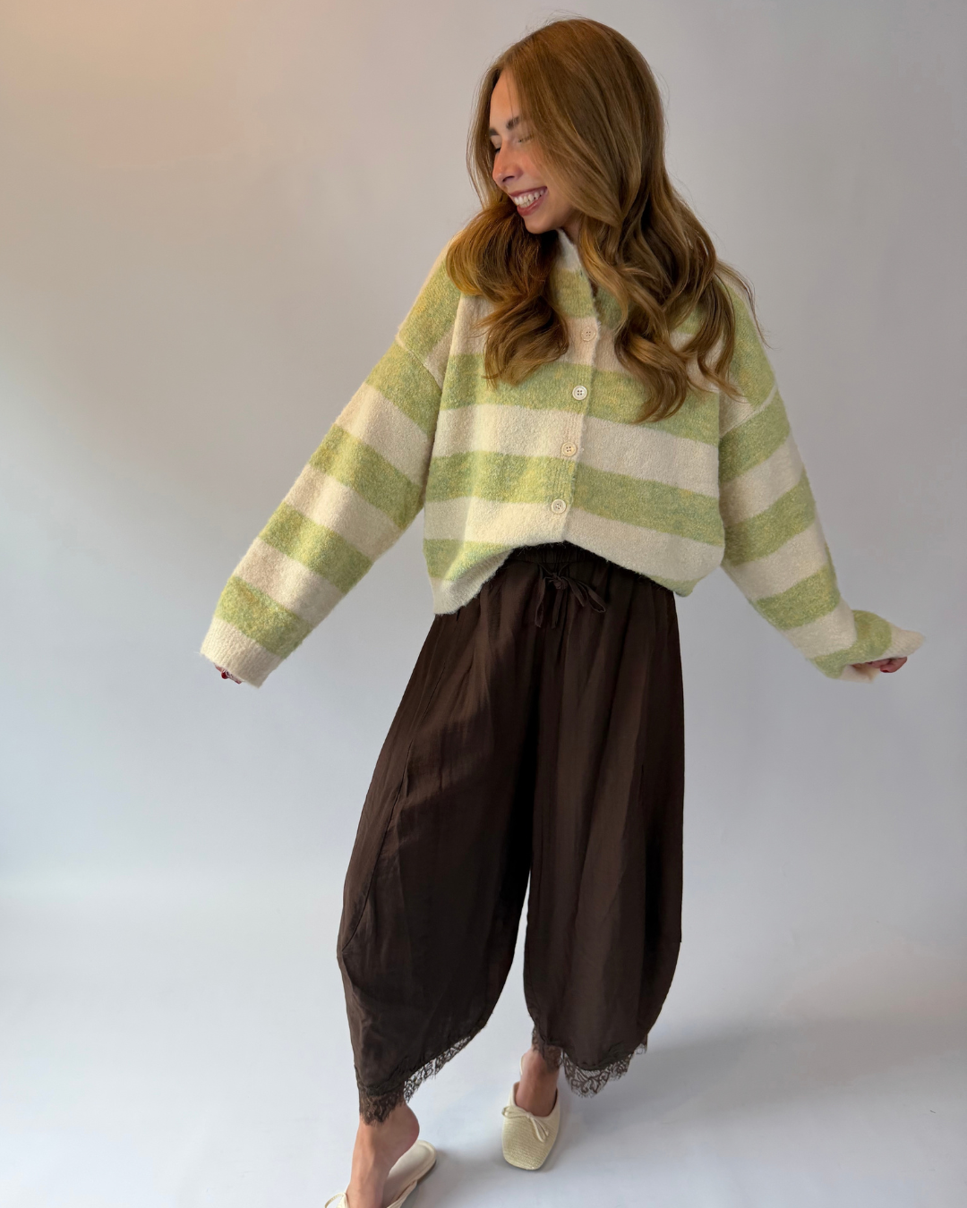 A woman modeling a fuzzy knit cardigan with light green and cream horizontal stripes, paired with dark brown harem-style pants and beige slip-on shoes, against a plain light gray studio background.