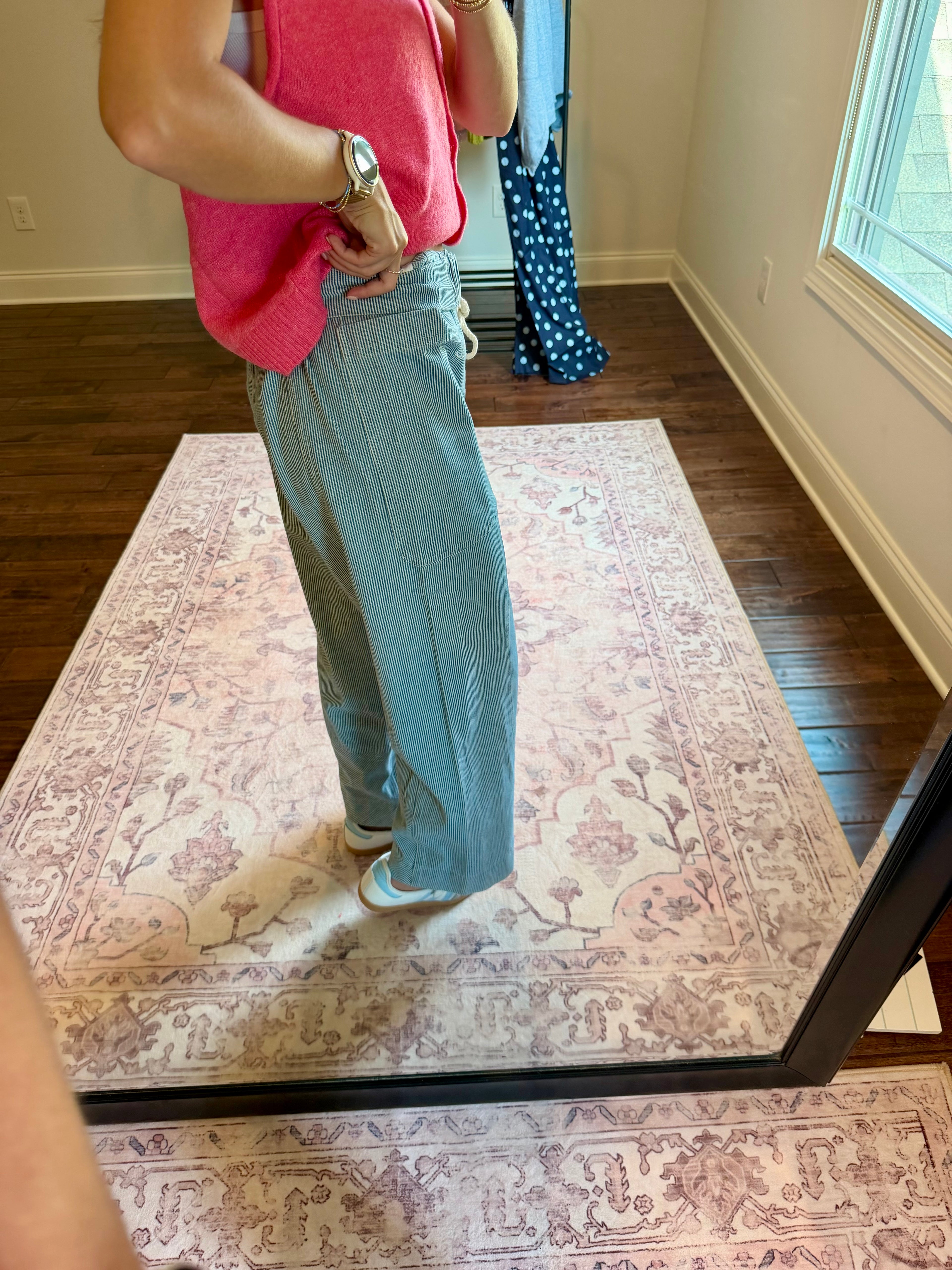 A person standing in a room taking a mirror selfie to show off a pair of wide-leg light blue jeans. The person is also wearing a bright pink sleeveless top and white sneakers.