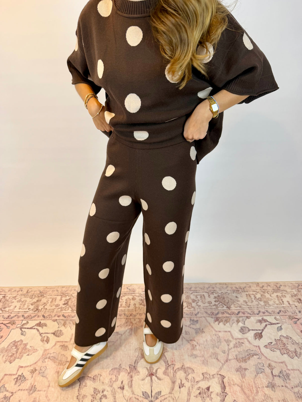 A woman wearing a matching brown knit two-piece set with large white polka dots, consisting of a short-sleeve top and wide-leg pants, paired with white sneakers, standing on a patterned rug.