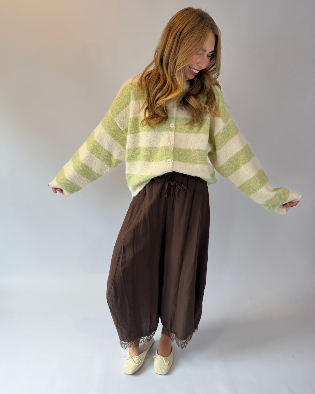 A woman modeling a light green and cream striped fuzzy cardigan, paired with dark brown wide-leg harem pants and cream-colored shoes, set against a plain light gray studio background.