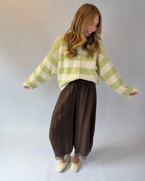 A woman modeling a light green and cream striped fuzzy cardigan, paired with dark brown wide-leg harem pants and cream-colored shoes, set against a plain light gray studio background.