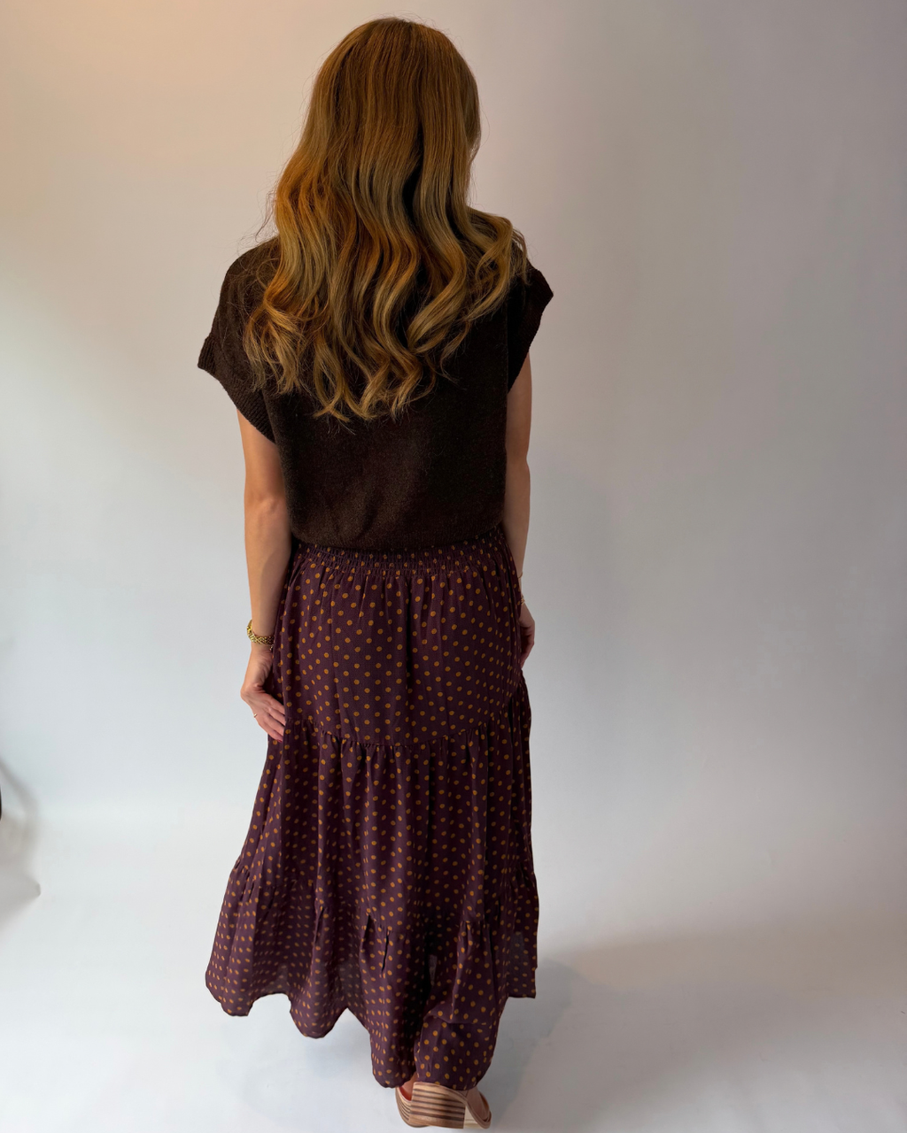 A woman viewed from behind wearing a dark brown short-sleeve top and a dark red tiered skirt with a small yellow pattern, standing against a plain light gray studio background.