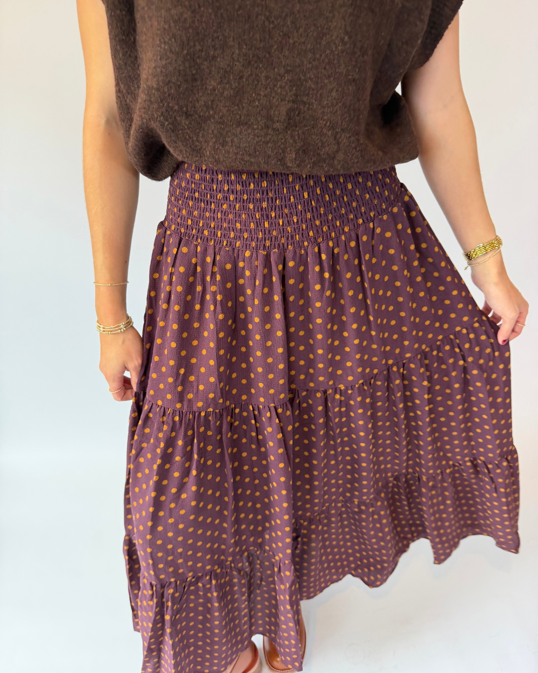 A person wearing a brown smocked-ruffle tiered skirt with small orange polka dots, paired with a brown knit top, shown from the waist down against a plain white background.