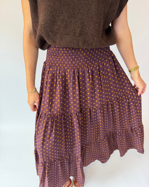 A person wearing a brown smocked-ruffle tiered skirt with small orange polka dots, paired with a brown knit top, shown from the waist down against a plain white background.
