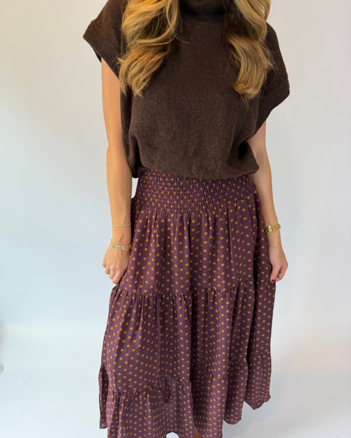 A woman wearing a dark brown short-sleeve knit top and a matching dark brown tiered skirt with small orange polka dots, standing against a plain light background.