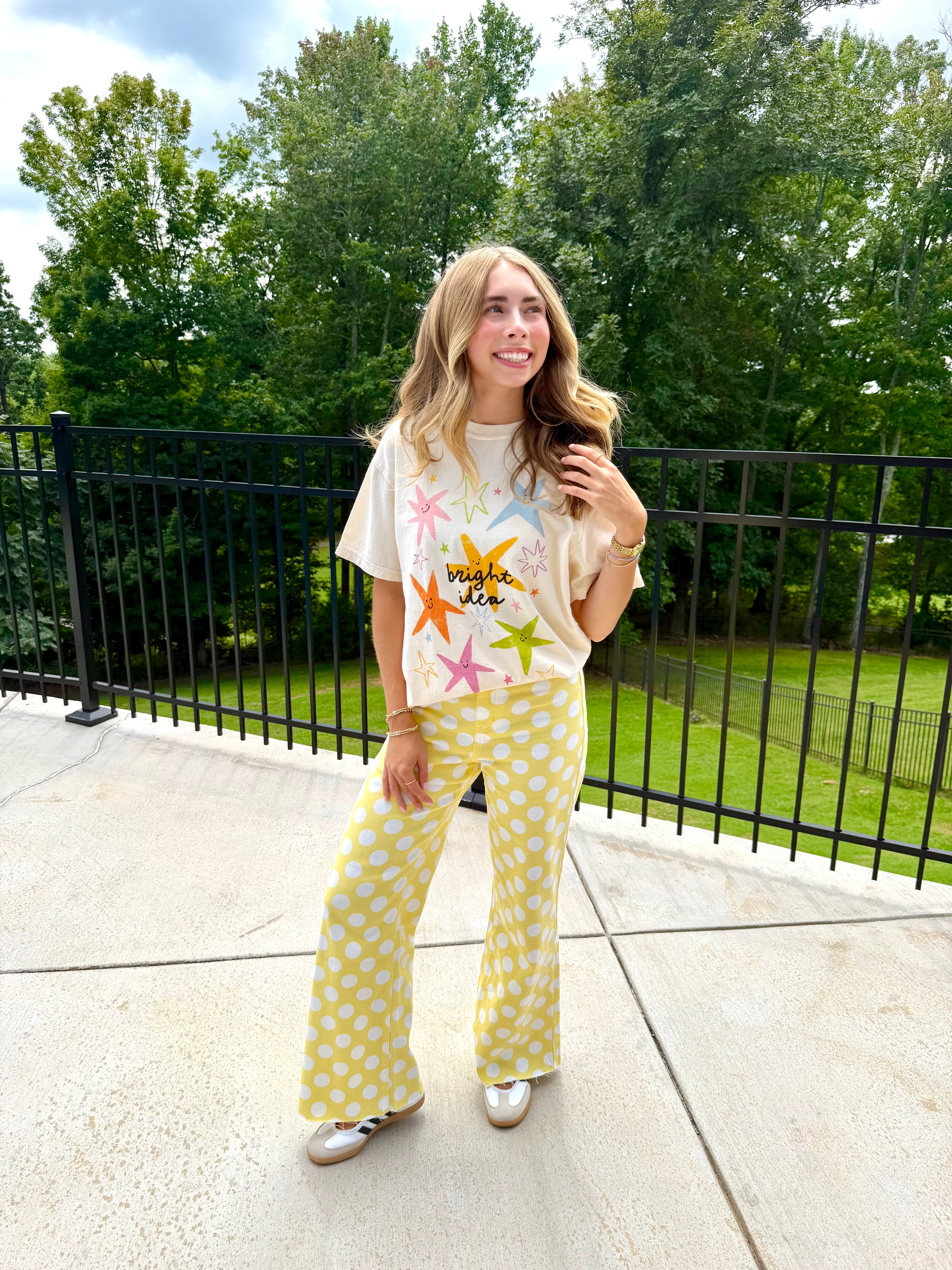 A woman stands outdoors on a concrete patio wearing a cream graphic t-shirt with colorful starfish prints and matching yellow-and-white polka dot wide-leg pants. Trees and a black metal fence are visible in the background.