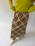 A person wearing a brown and beige plaid maxi skirt with a bright yellow-green sweater and white sneakers, standing against a plain white studio background.
