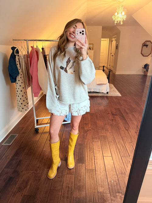 Women taking a mirror selfie in a room with wooden flooring and a clothing rack. Women is wearing a duck sweater, white shorts, and yellow boots. 