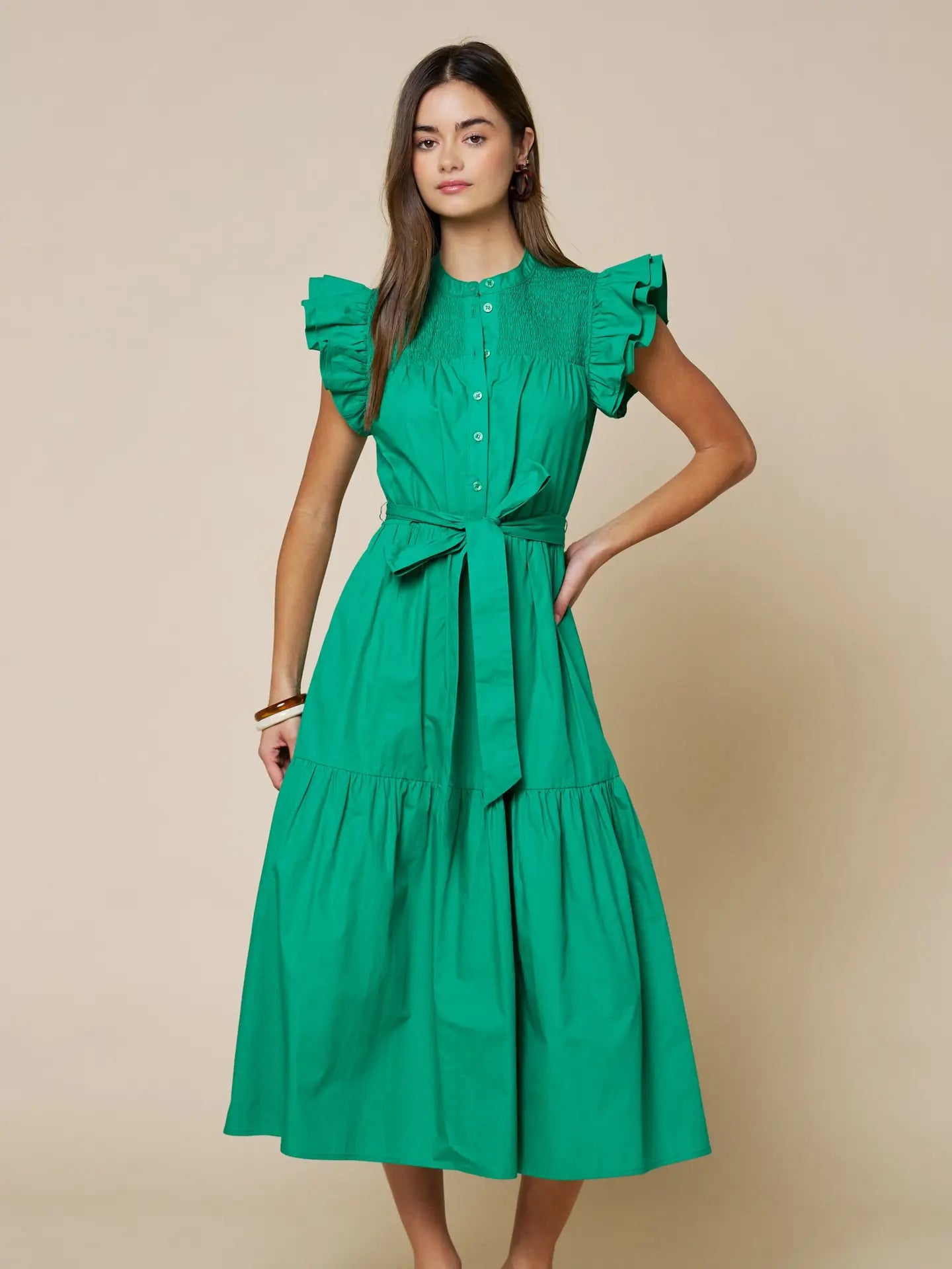 A woman modeling a bright green midi dress with ruffled cap sleeves, a buttoned bodice, a matching waist tie, and a tiered skirt, set against a solid beige studio background.