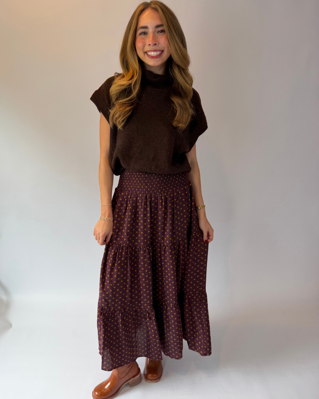 A woman modeling a dark brown short-sleeve mock neck top and a dark red tiered maxi skirt with a small white polka dot pattern, paired with brown leather shoes, against a plain light gray studio background.
