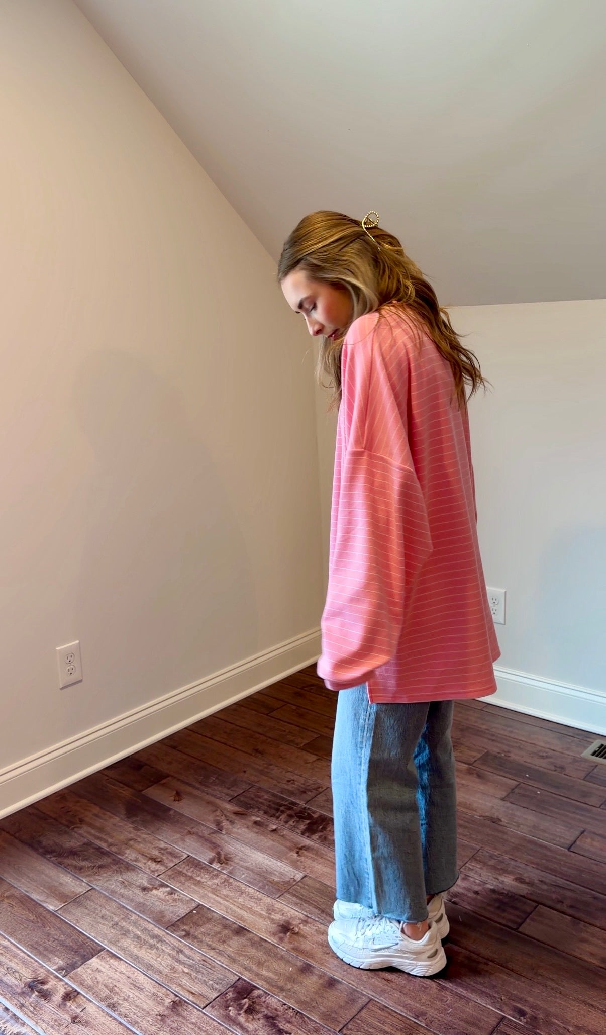 Woman wearing a pink sweater and blue jeans standing in a room with wooden flooring.