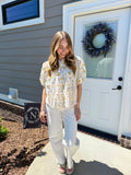 Woman standing outside a house with a wreath on the door, wearing a floral blouse and white pants.