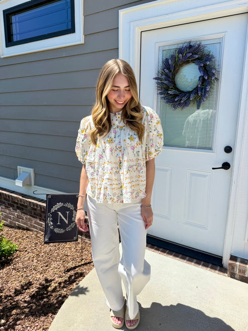 Woman standing outside a house with a wreath on the door, wearing a floral blouse and white pants.