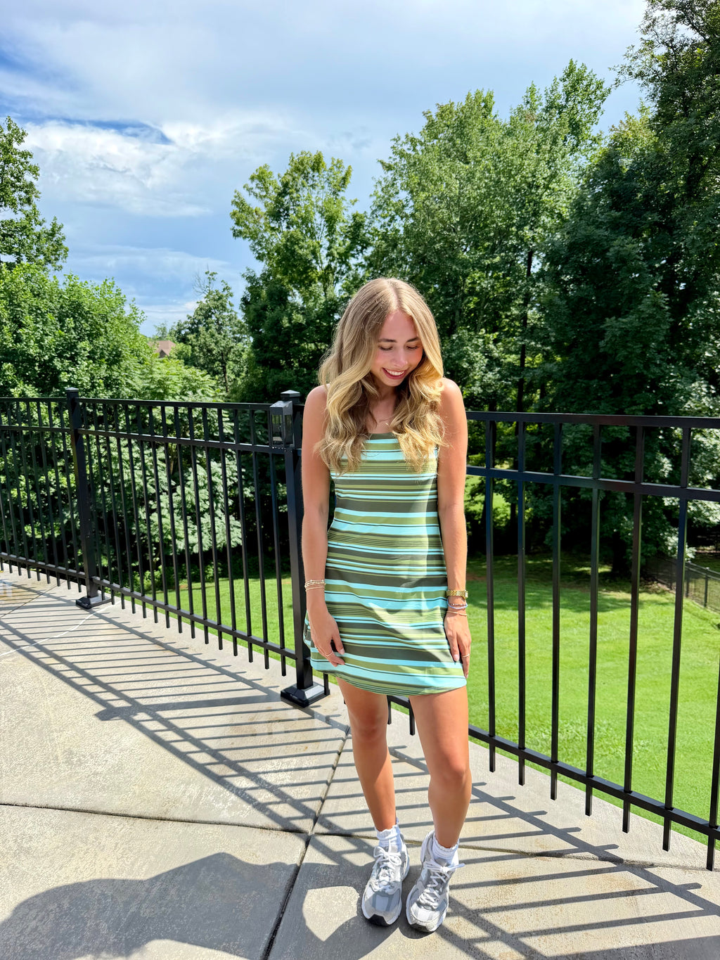 A woman stands on an outdoor balcony wearing a sleeveless, mini-length dress with horizontal stripes in mint green, teal, and white. She is wearing white athletic sneakers and standing in front of a black metal railing with green trees and grass in the background.