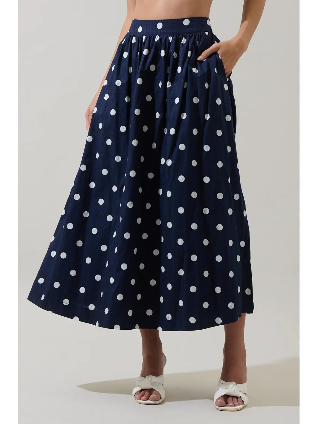 A navy blue midi skirt with white polka dots, worn by a model with white slide sandals against a light gray studio background.