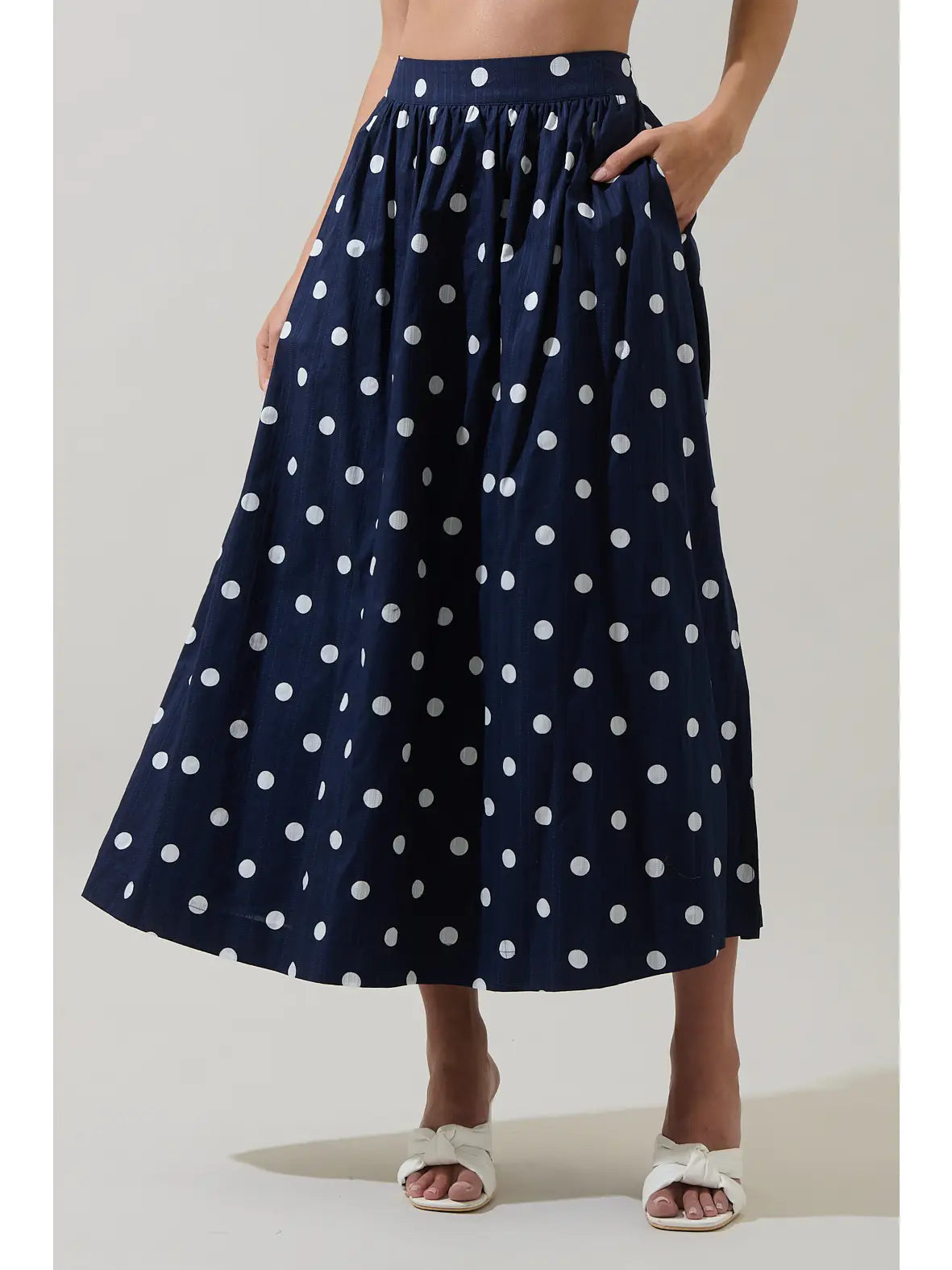 A navy blue midi skirt with white polka dots, worn by a model with white slide sandals against a light gray studio background.