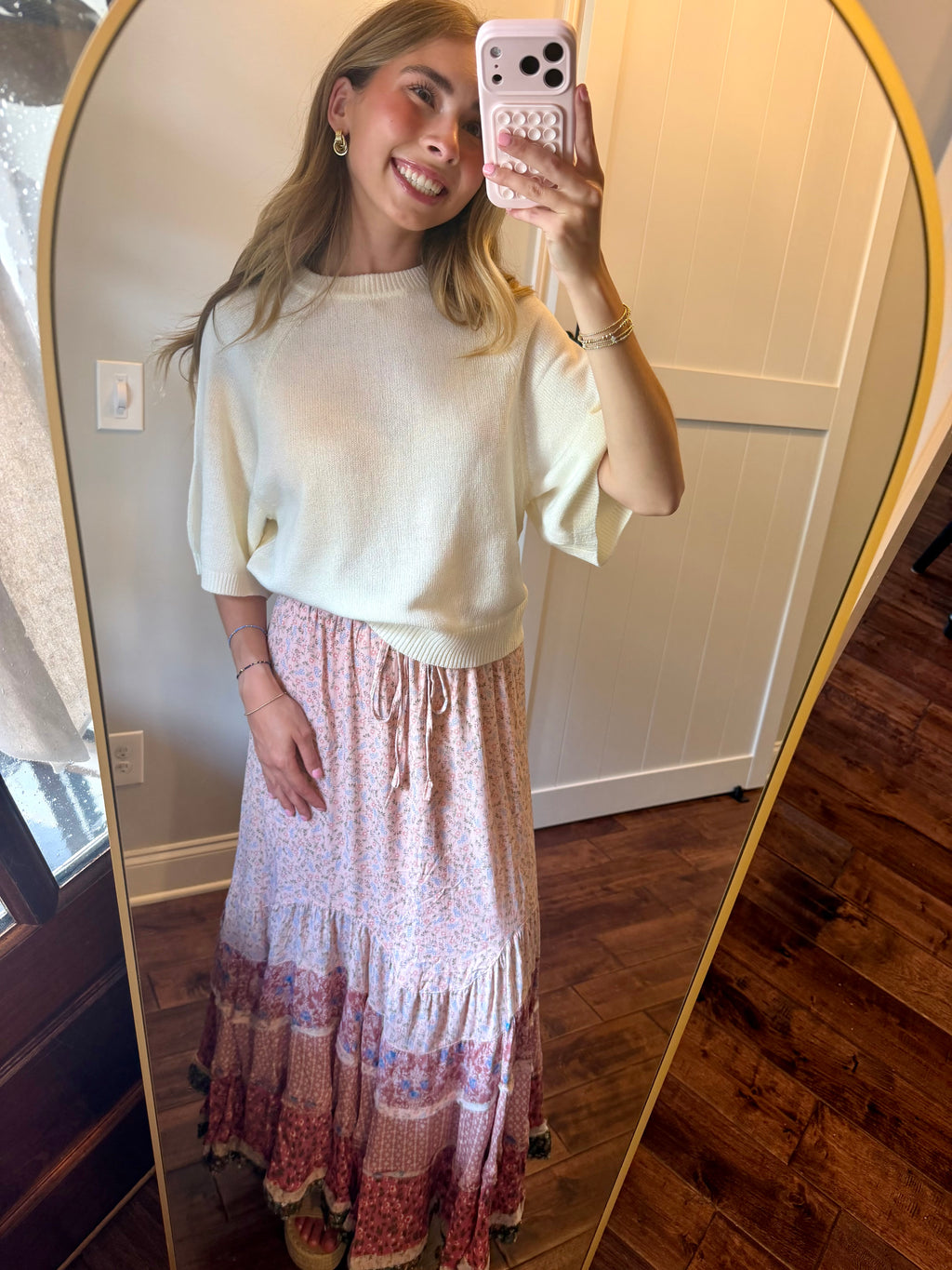 Woman taking a mirror selfie wearing a white top and pink floral skirt.