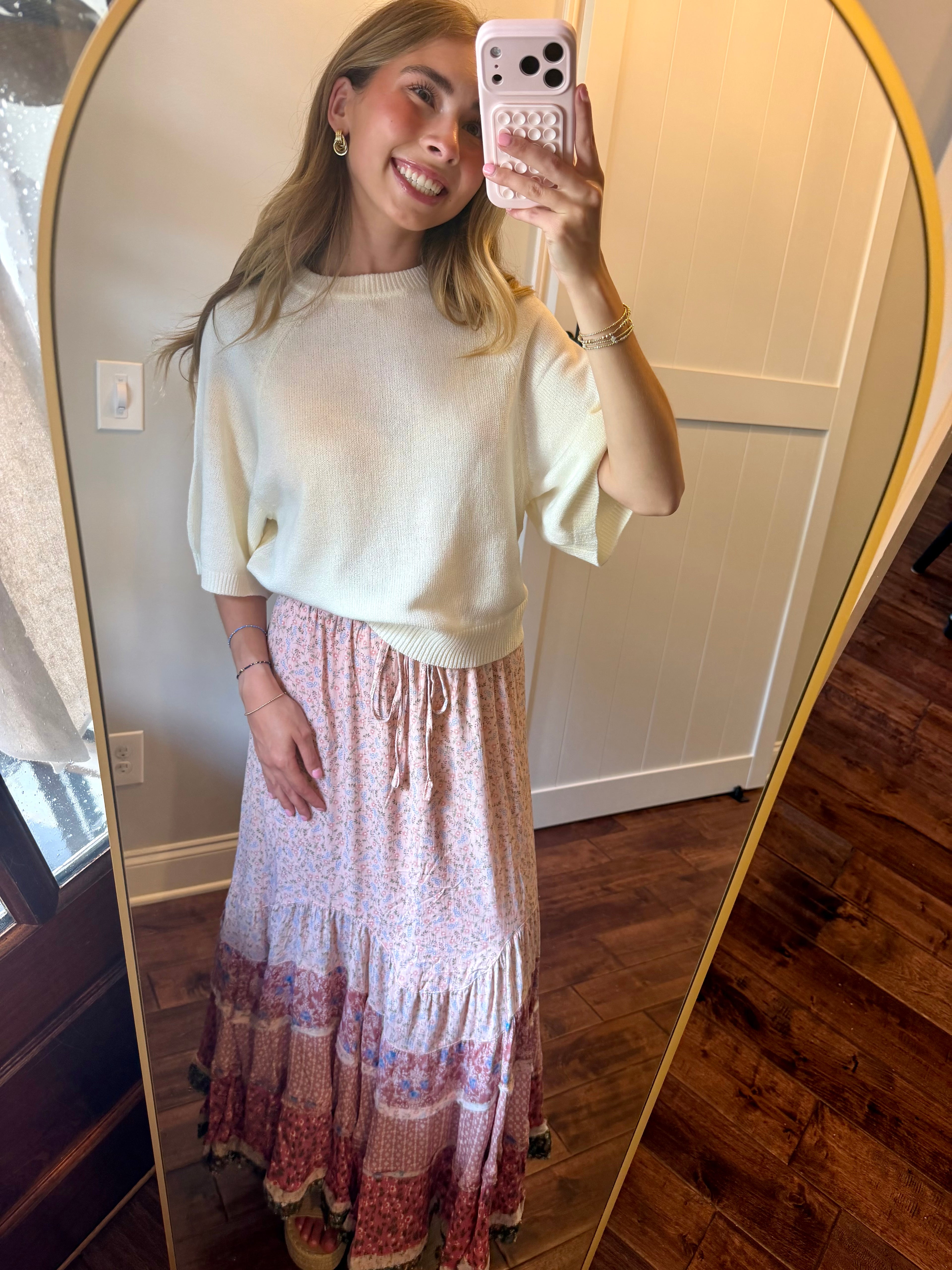 Woman taking a mirror selfie wearing a white top and pink floral skirt.