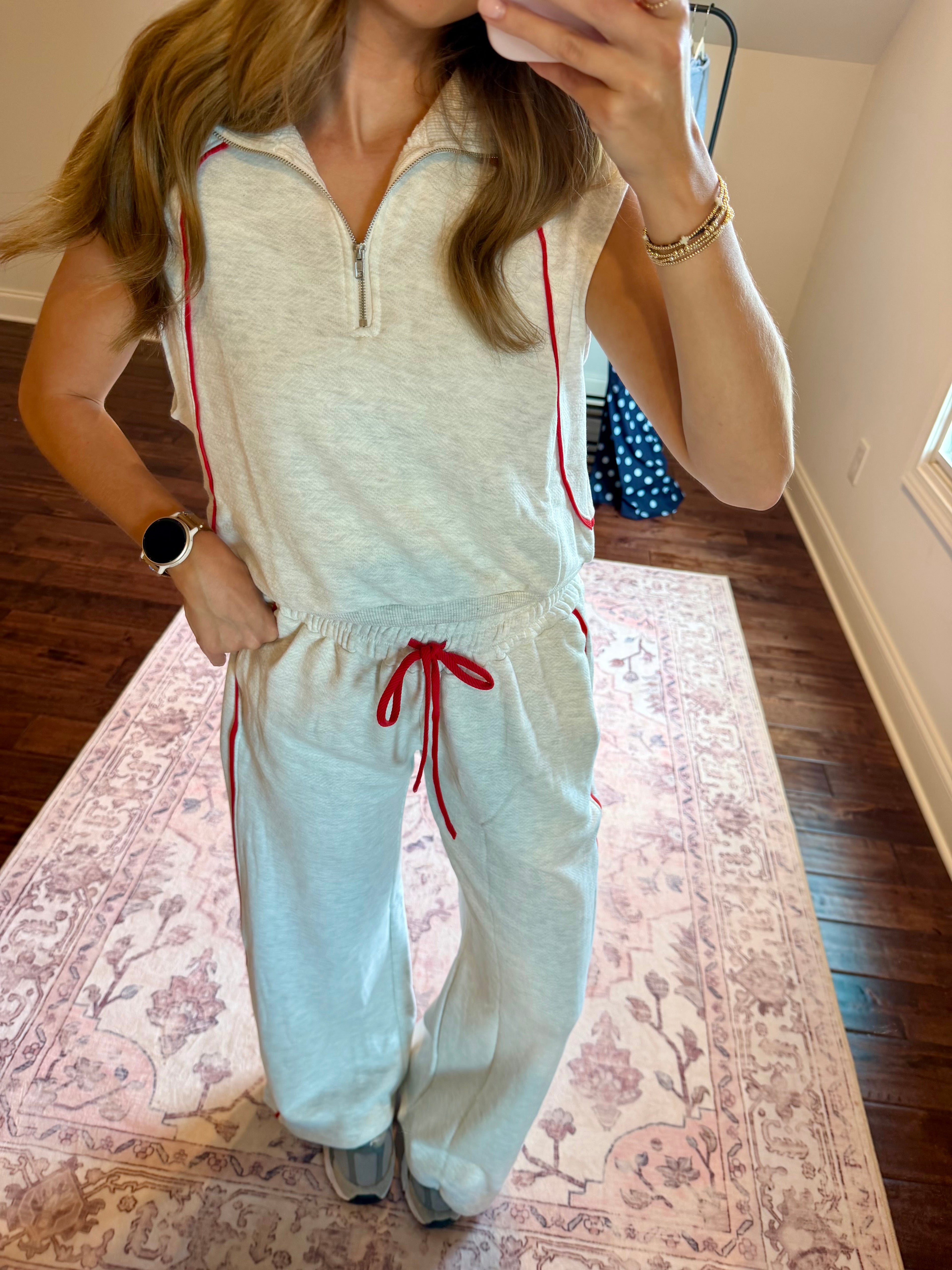 A woman takes a mirror selfie wearing a light gray two-piece tracksuit with red accents, featuring a sleeveless quarter-zip top and matching sweatpants with a red drawstring.