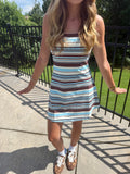 A woman wearing a sleeveless A-line mini dress with horizontal brown, light blue, and white stripes, standing on an outdoor concrete walkway next to a black metal fence.