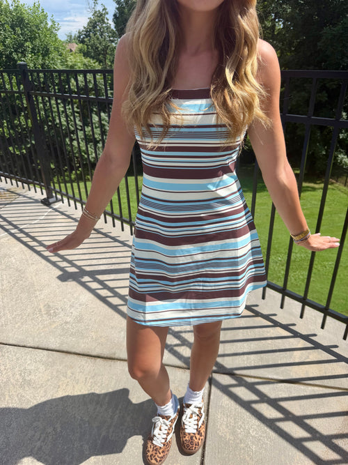 A woman wearing a sleeveless A-line mini dress with horizontal brown, light blue, and white stripes, standing on an outdoor concrete walkway next to a black metal fence.