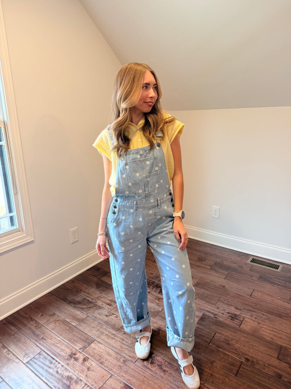 Woman wearing a yellow top and light blue overalls standing in a room with wooden flooring.