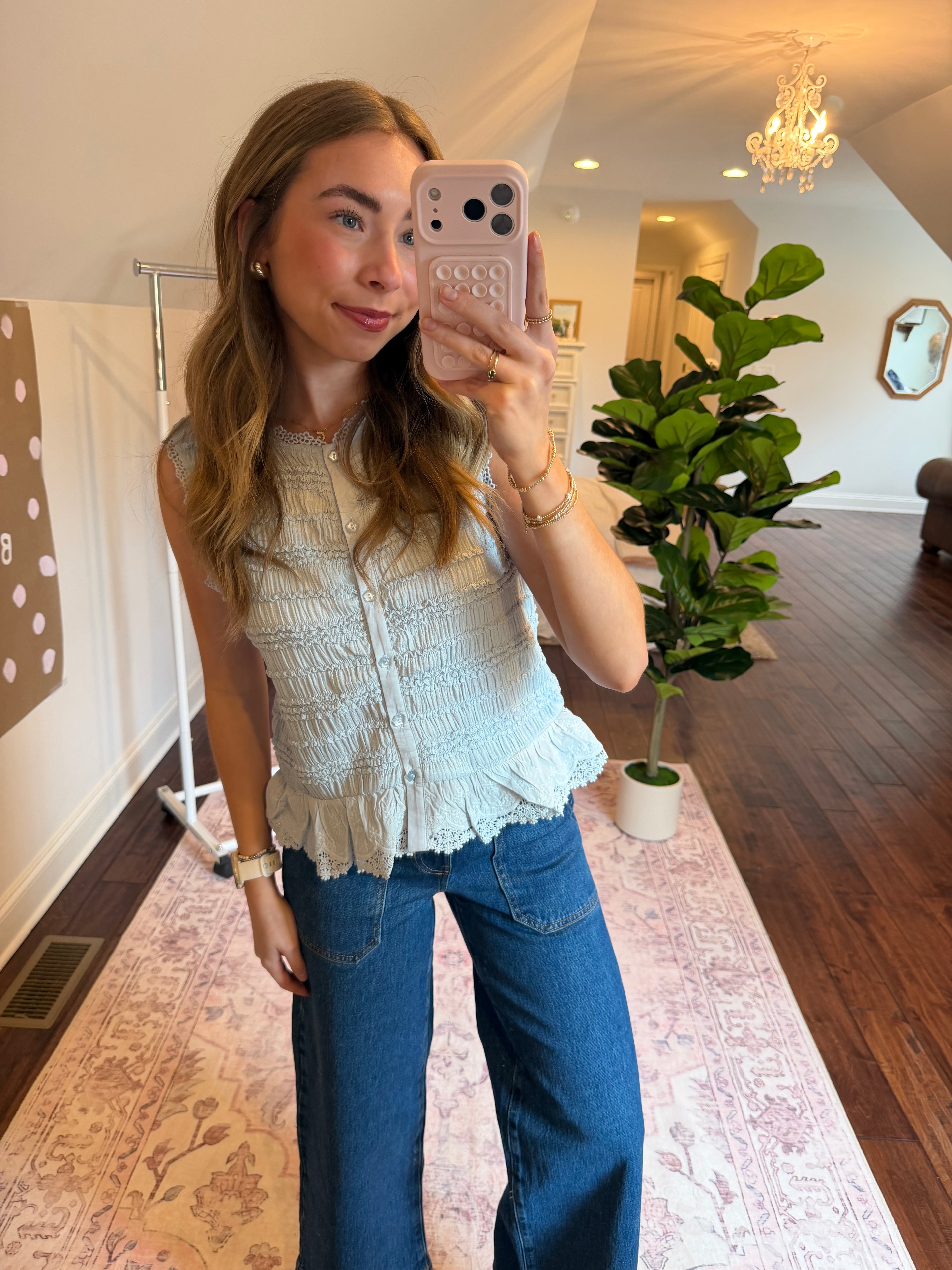 Woman taking a selfie in a room with a plant and decorative items. Wearing a baby blue tank top.
