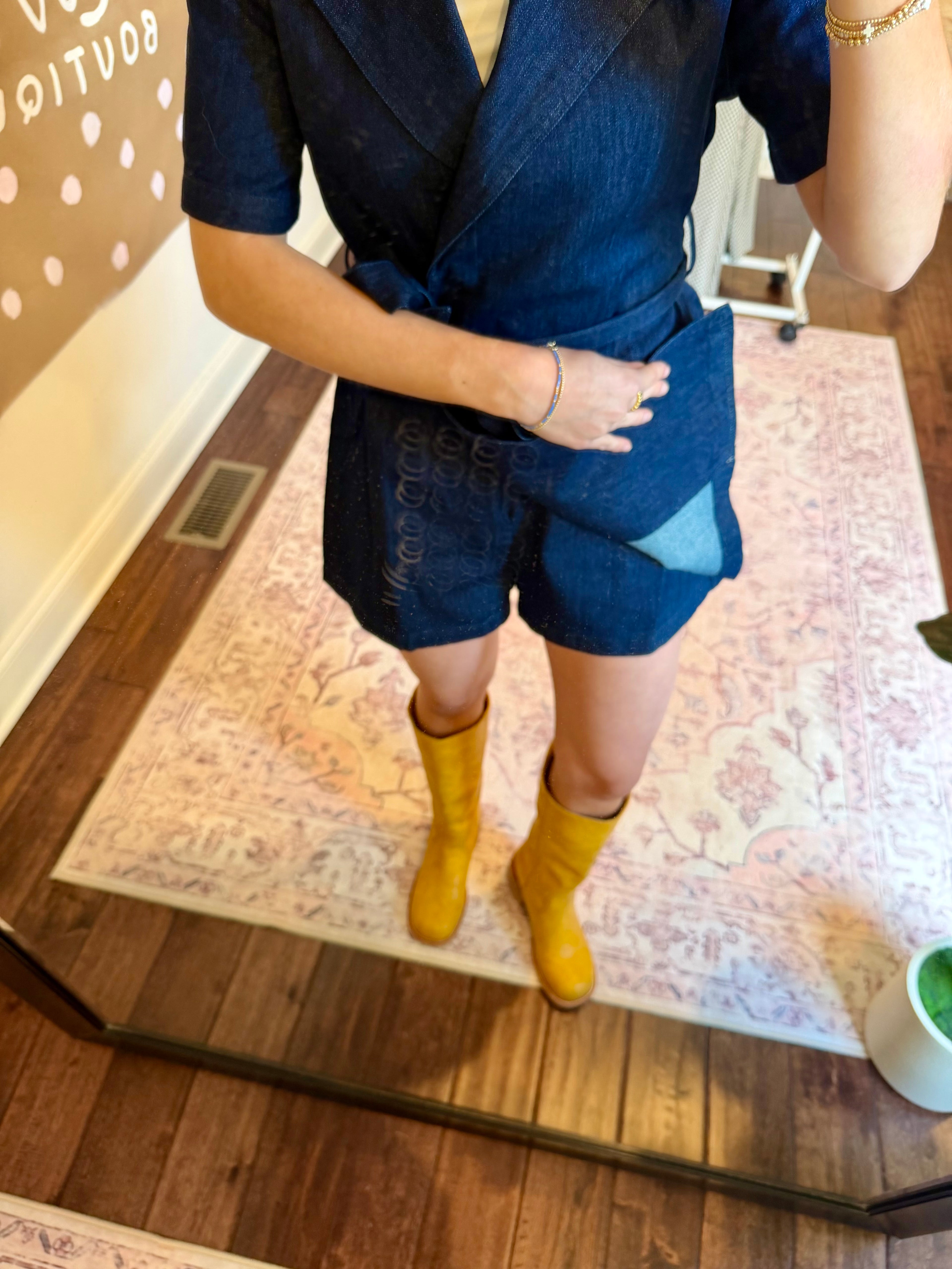 Woman taking a mirror selfie wearing a blue dress and yellow boots in a room with a plant and decorative wall art.
