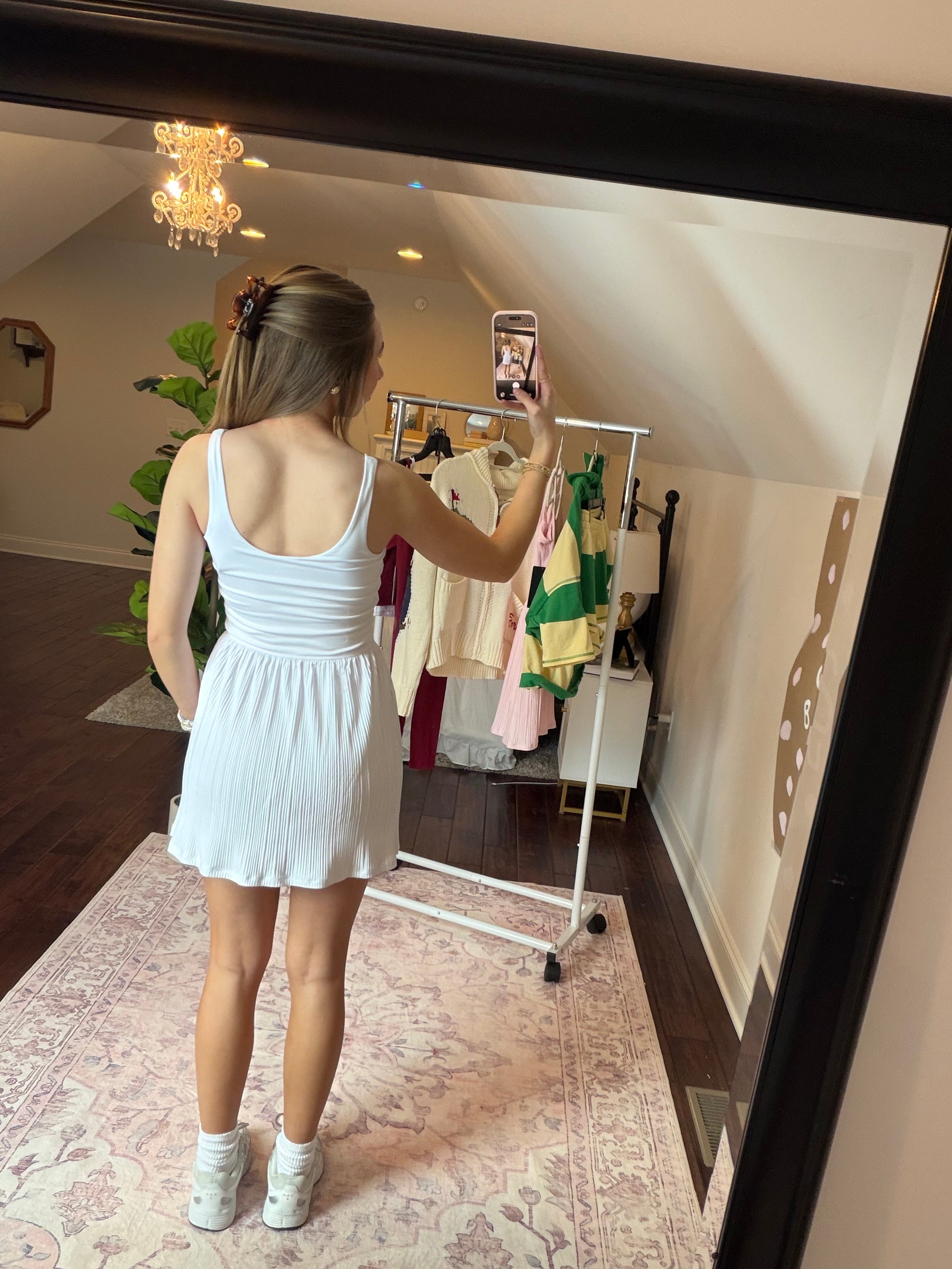 Person taking a mirror selfie wearing a white dress in a room with a clothing rack.