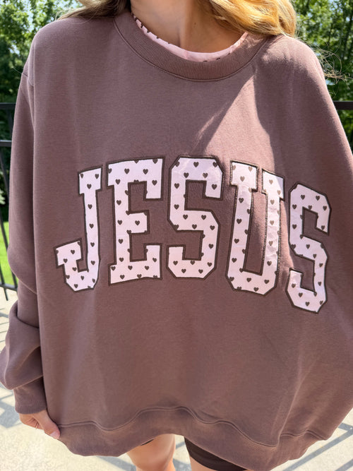 A person wearing an oversized brown sweatshirt with the word 'JESUS' printed across the front in large white letters with small pink hearts and dark outlines, photographed outdoors.