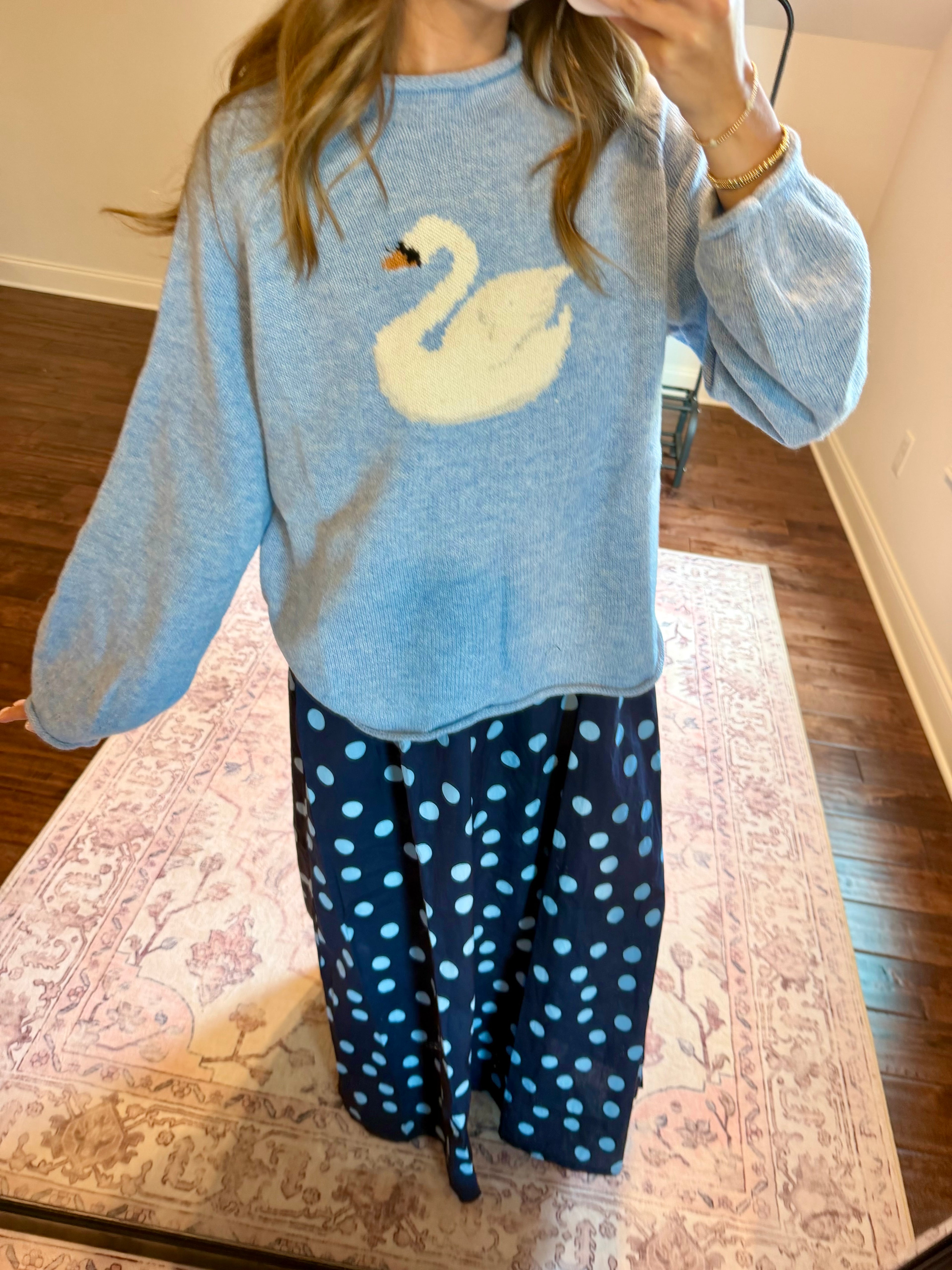 A person wearing a light blue knit sweater with a large white swan motif on the front, paired with navy pants covered in light blue polka dots, standing on a patterned rug in an indoor room.