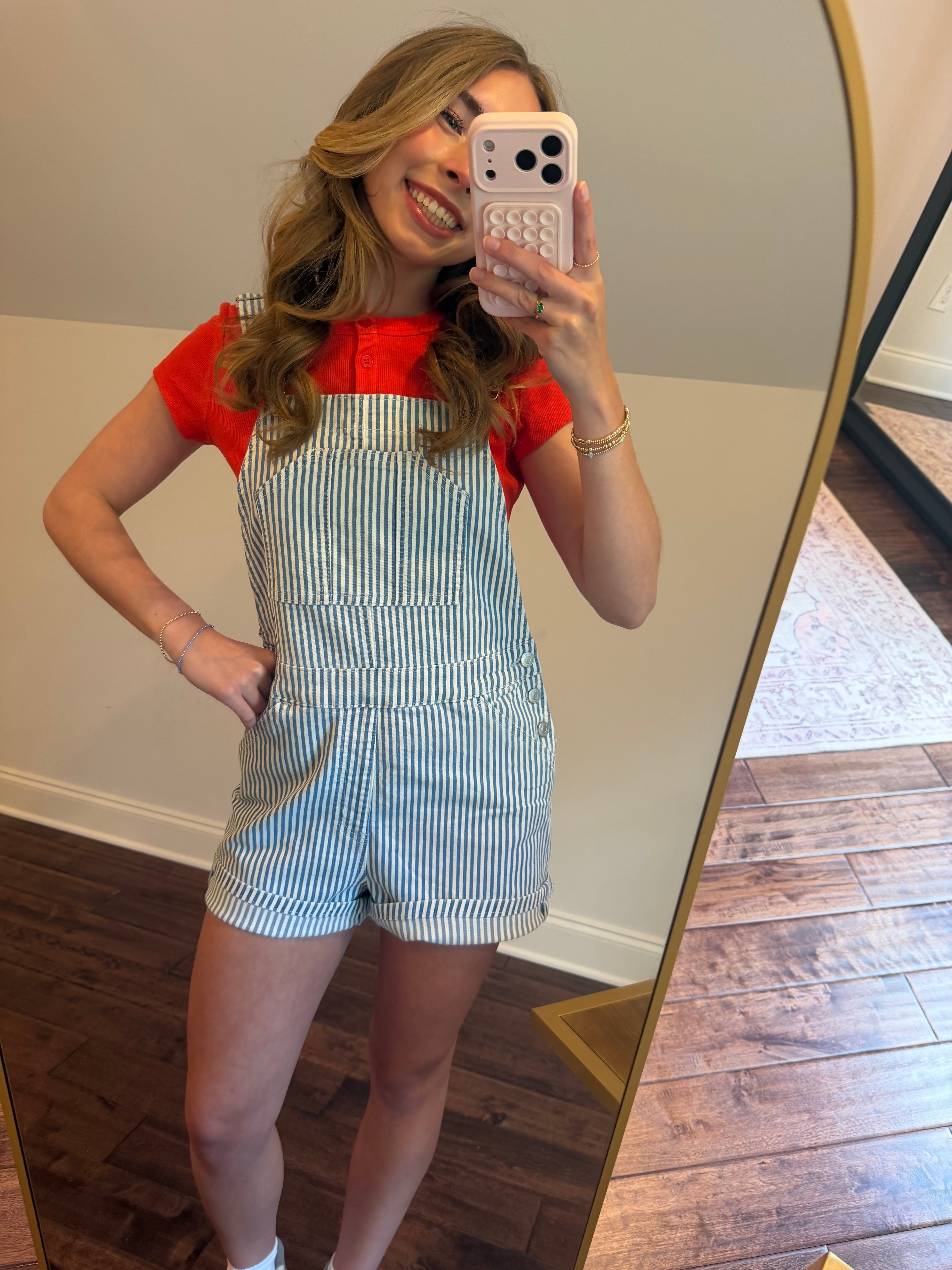 Woman taking a mirror selfie wearing a red top and striped overalls.