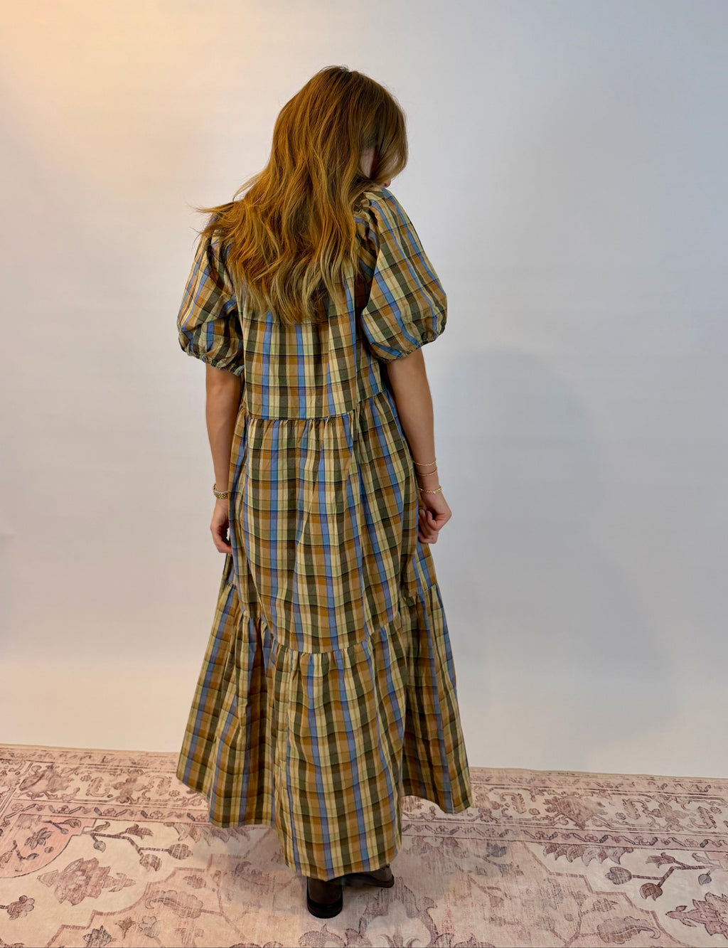 A woman viewed from behind wearing a yellow, blue, and brown plaid midi dress with puff sleeves, standing on a patterned rug against a plain white wall.