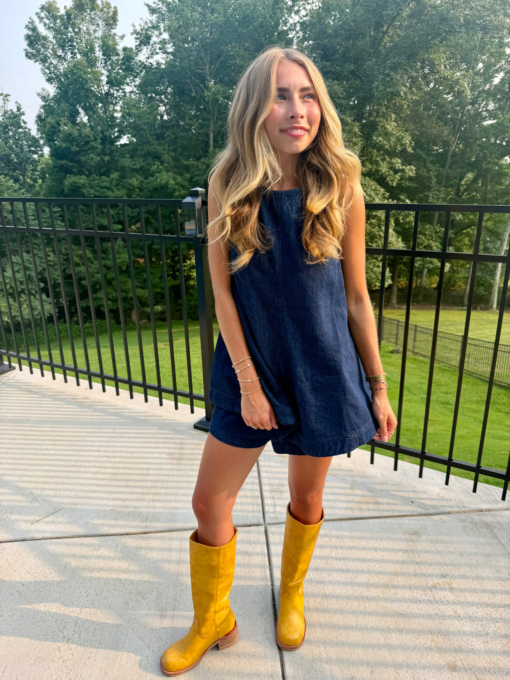 A woman stands outdoors on a concrete patio wearing a sleeveless dark blue denim romper and bright yellow knee-high boots, with a black metal fence and green trees in the background.