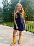 A woman stands outdoors on a concrete patio wearing a sleeveless dark blue denim romper and bright yellow knee-high boots, with a black metal fence and green trees in the background.