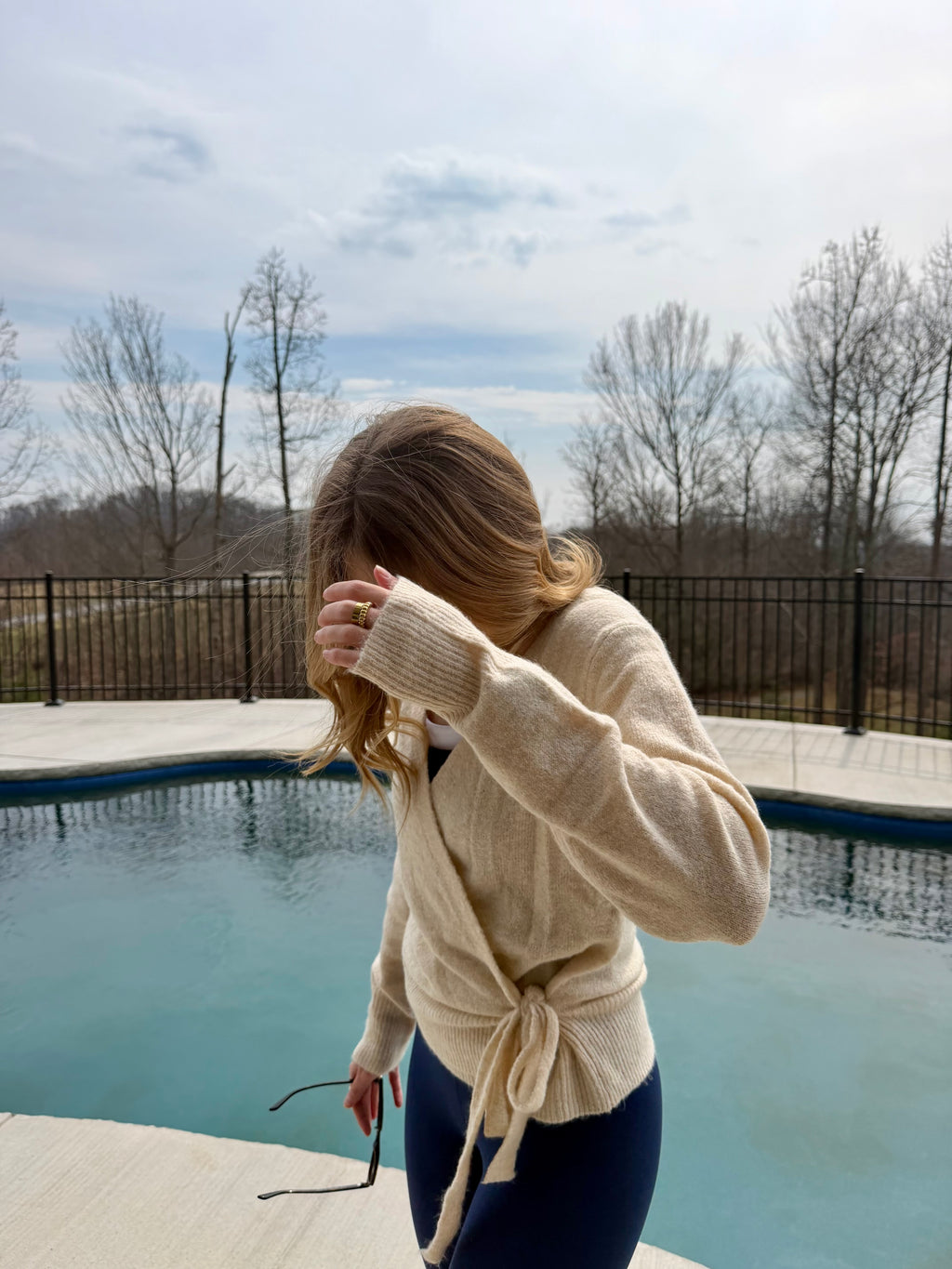 Woman in a beige cardigan standing by a pool with trees and a cloudy sky in the background
